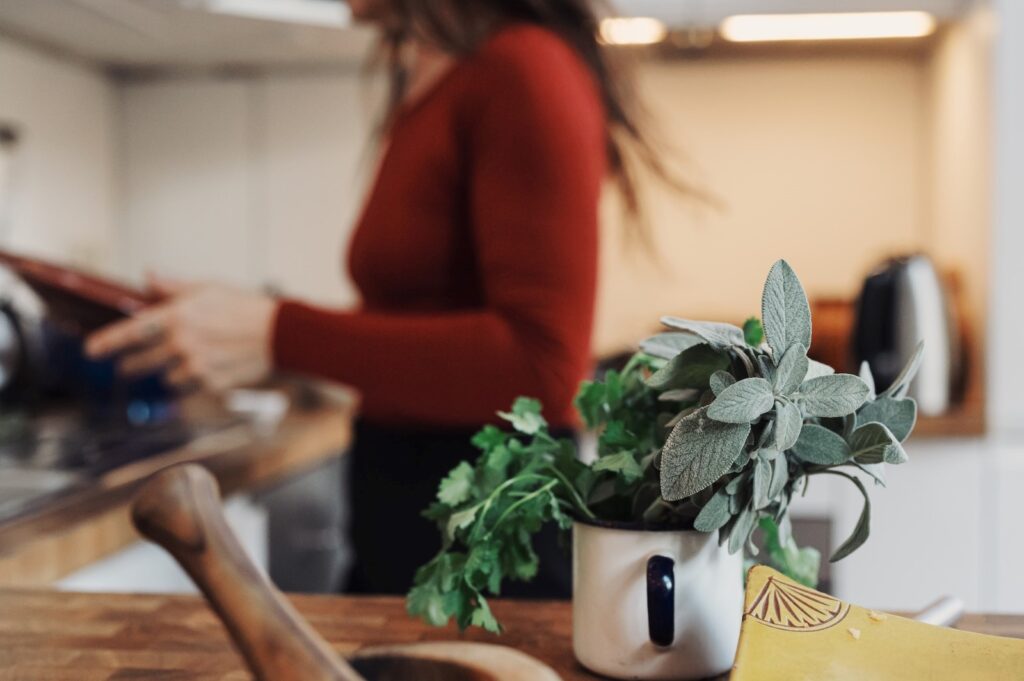A jar of fresh herbs in a mug on a counter with a woman wearing a red sweater behind it. 