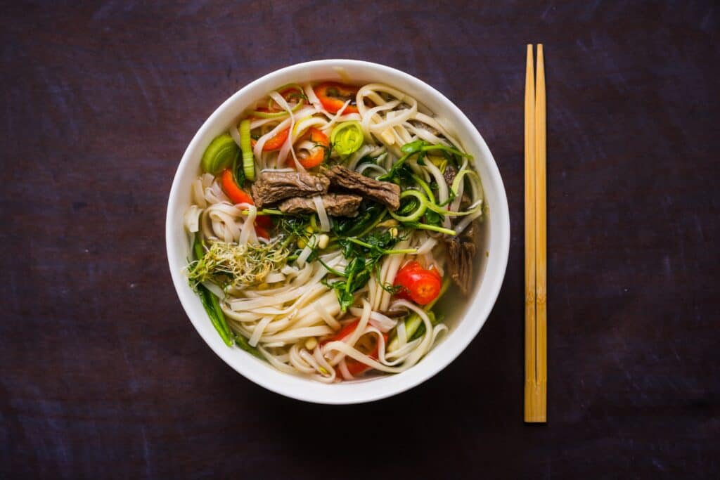 Rice noodles in a bowl with meat and vegetables and chopsticks on the side. 