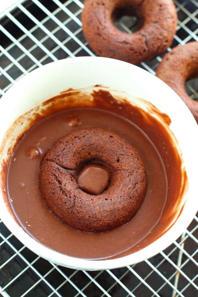 Baked gluten free chocolate donuts getting dipped in a chocolate glaze.