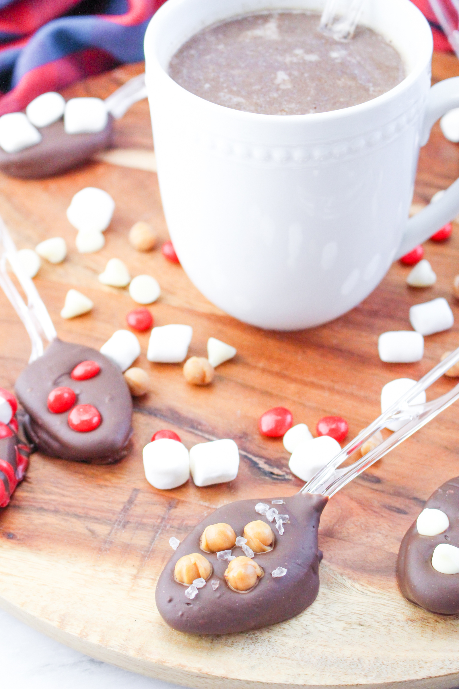 4 chocolate-dipped spoons with candy and marshmallows in the background and a white mug.