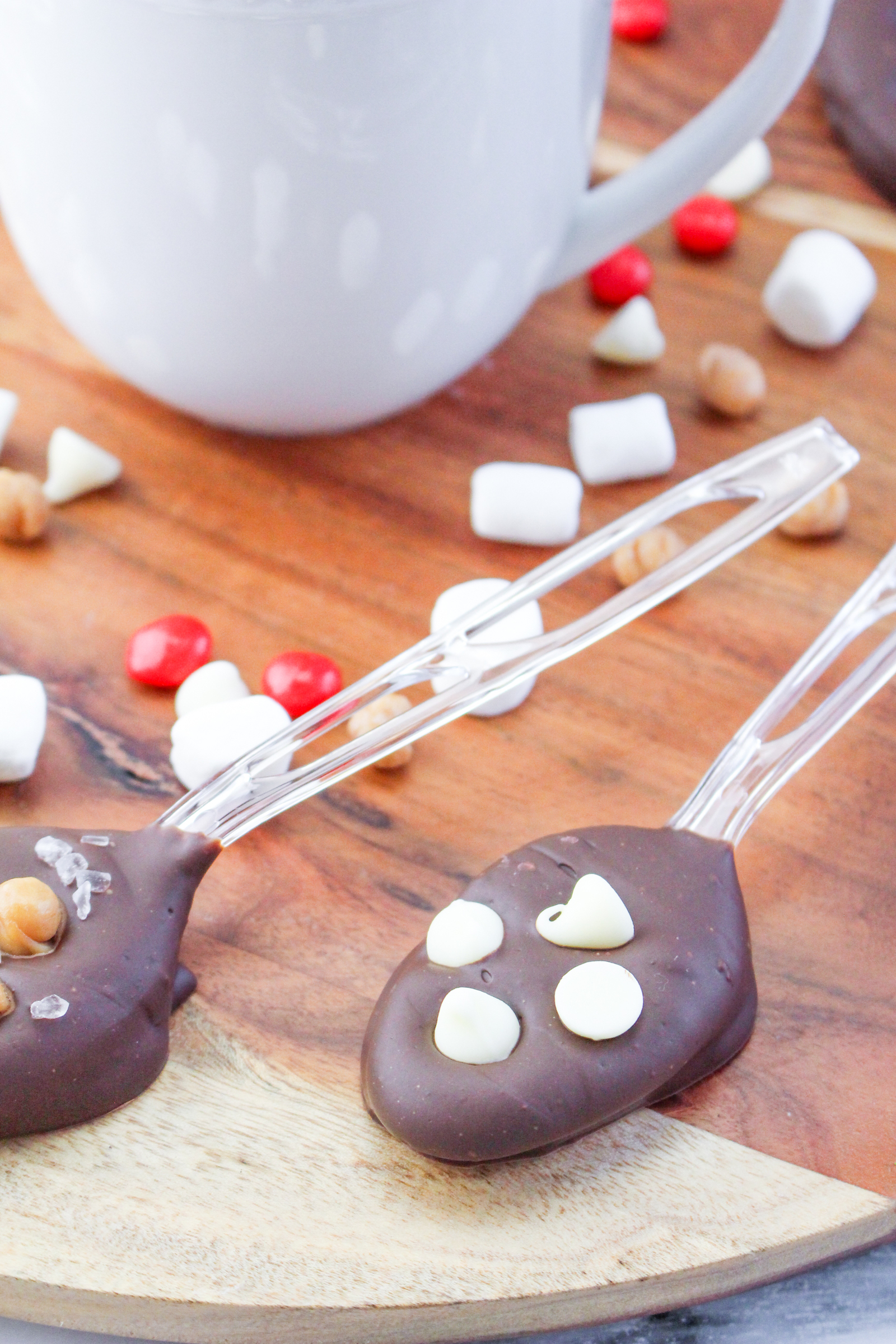Up close of two hot chocolate spoons on a wooden background with candy and marshmallows in the background.