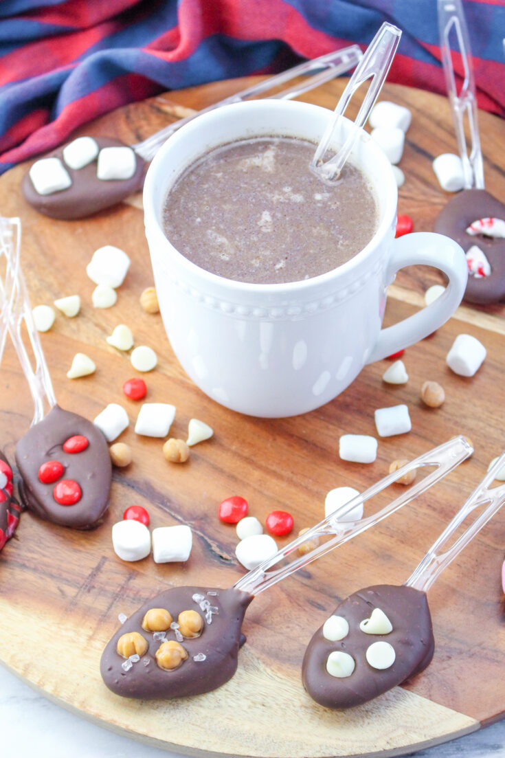 A white mug of hot chocolate with various diy hot chocolate spoons on a wooden background.