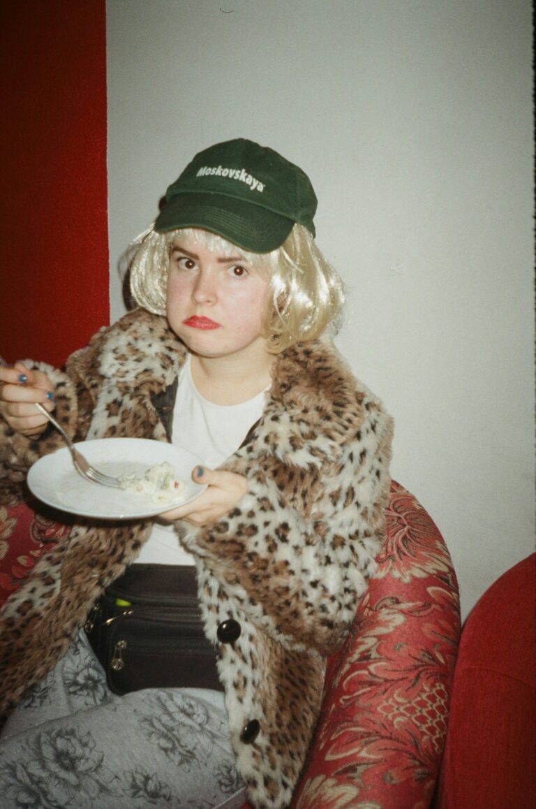 A kid dressed as a woman with blonde hair, a leopard coat, and a fanny pack is eating a plate of food.