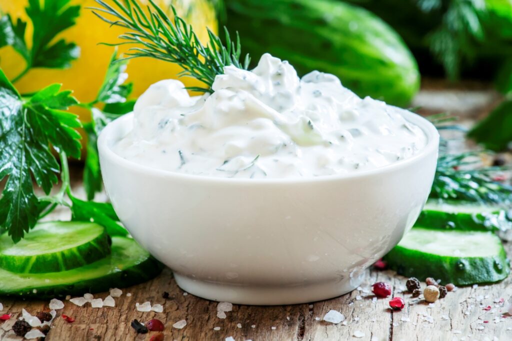 A white bowl of tzatziki sauce with vegetables behind it.