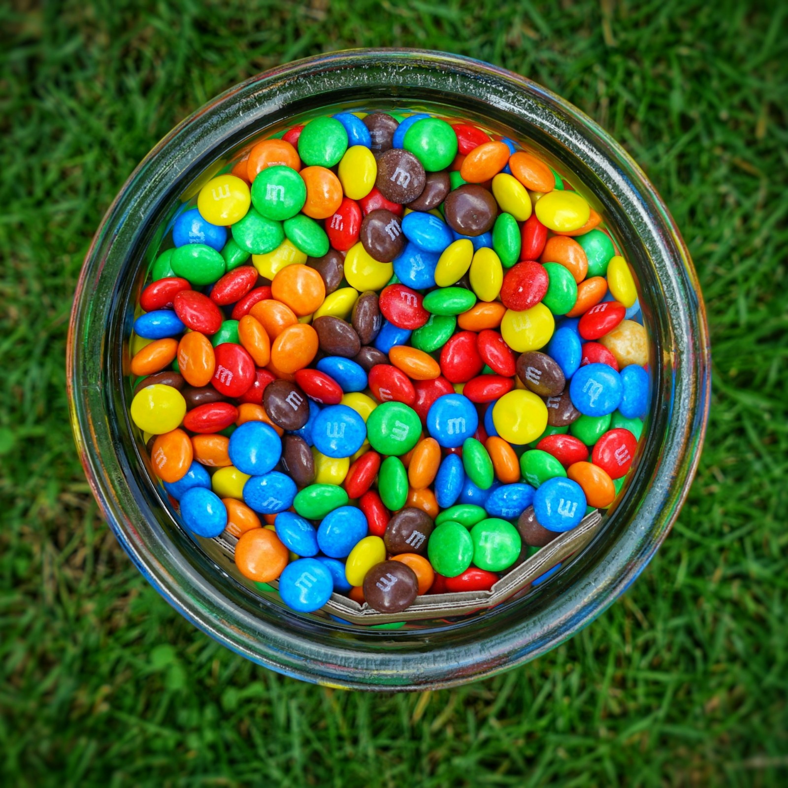 A glass jar filled with M&M's on green grass.