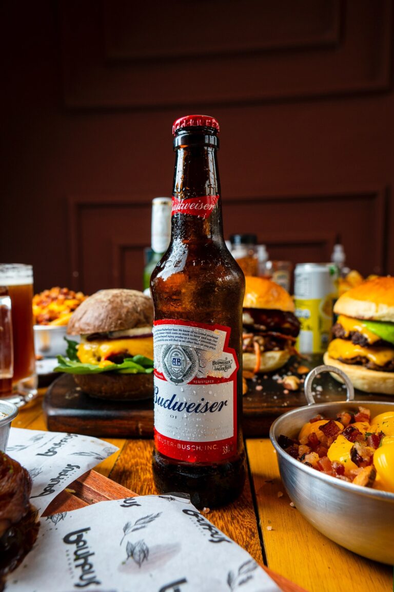A bottle of Budweiser surrounded by hamburgers and side dishes.