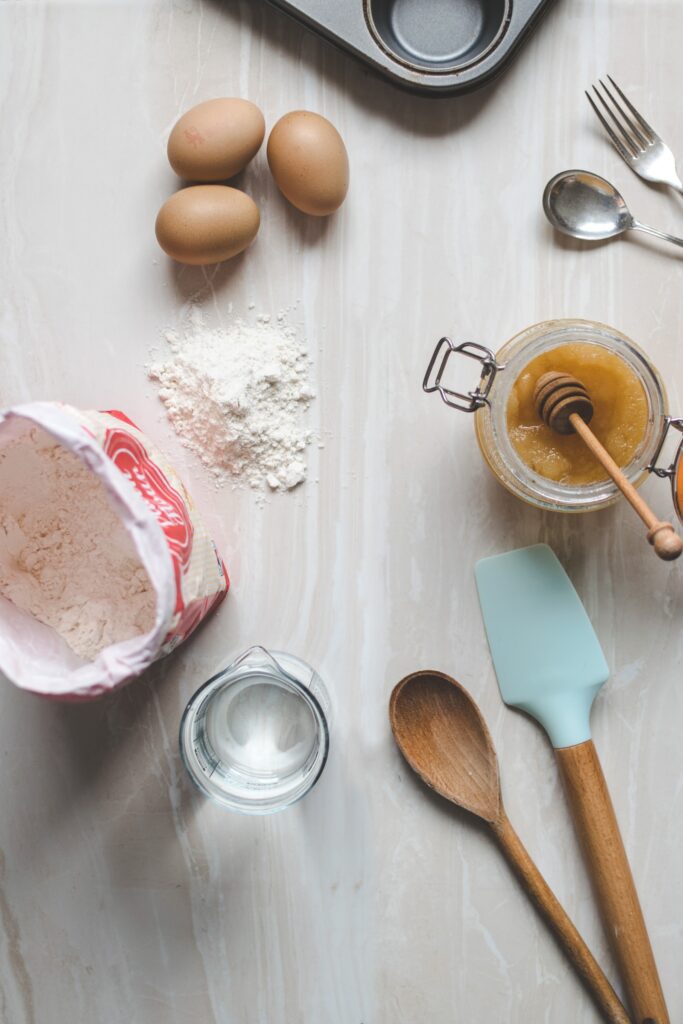 An overhead view of various baking ingredients: flour, honey, eggs, and measuring spoons.