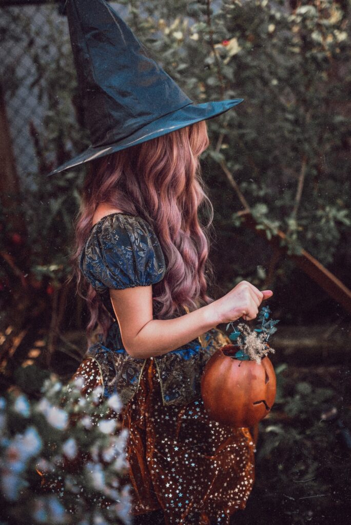 A young girl wearing a witch costume is carrying a pumpkin bucket.