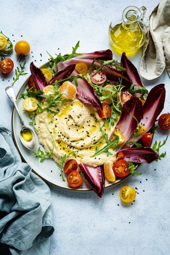 A bowl of hummus with vibrant vegetables like endive and sliced cherry tomatoes.