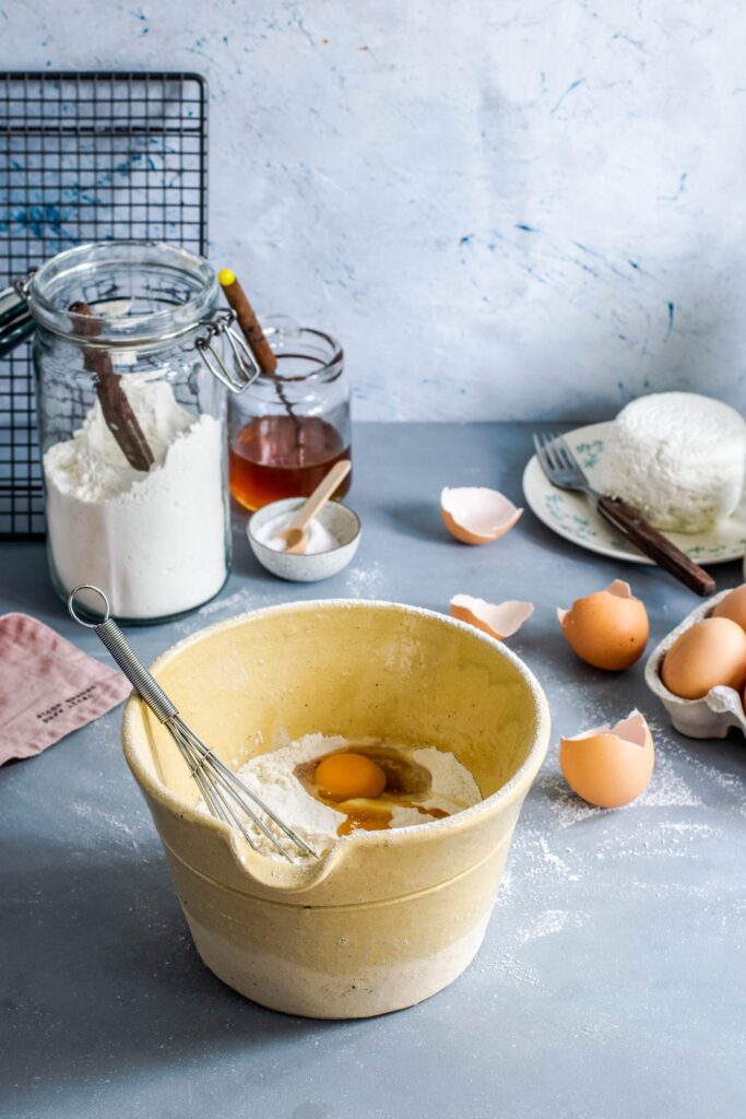 A yellow bowl filled with flour and eggs and a whisk with various baking ingredients in the background.