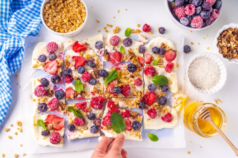 A sheet of Greek yogurt bark with assorted berries and mint and a hand grabbing one piece.