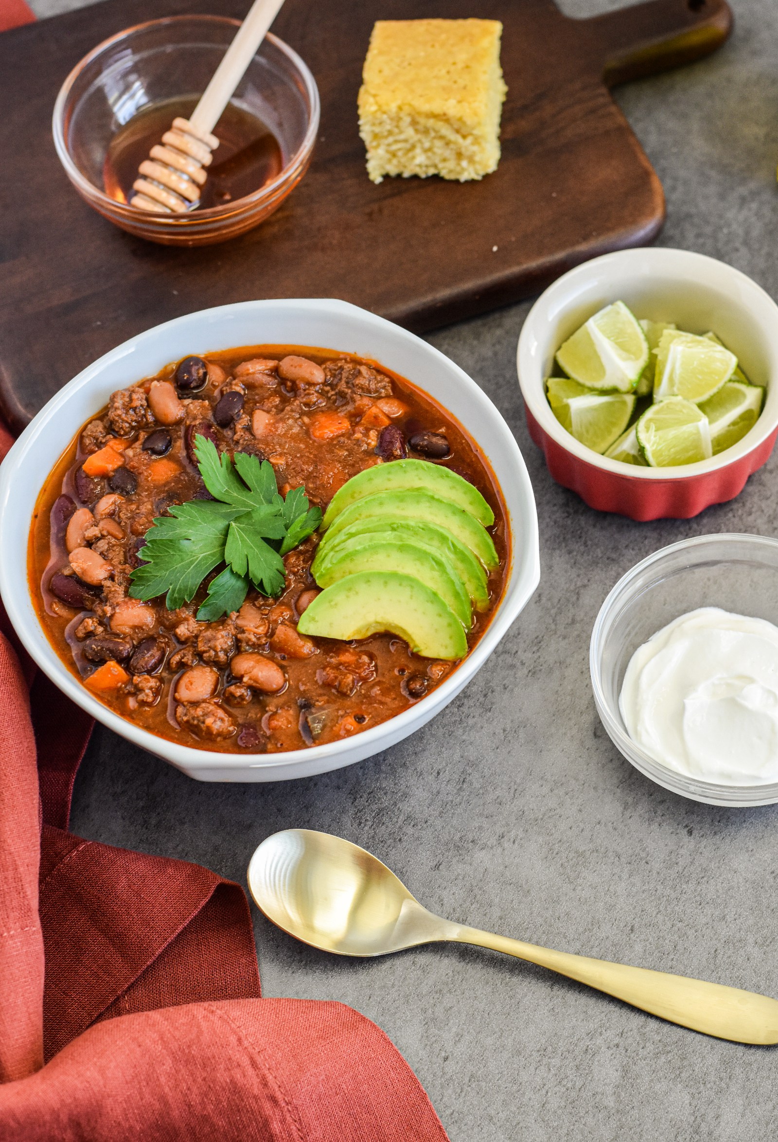 A white bowl filled with 3 bean chili and a bowl of sliced limes and a spoon.