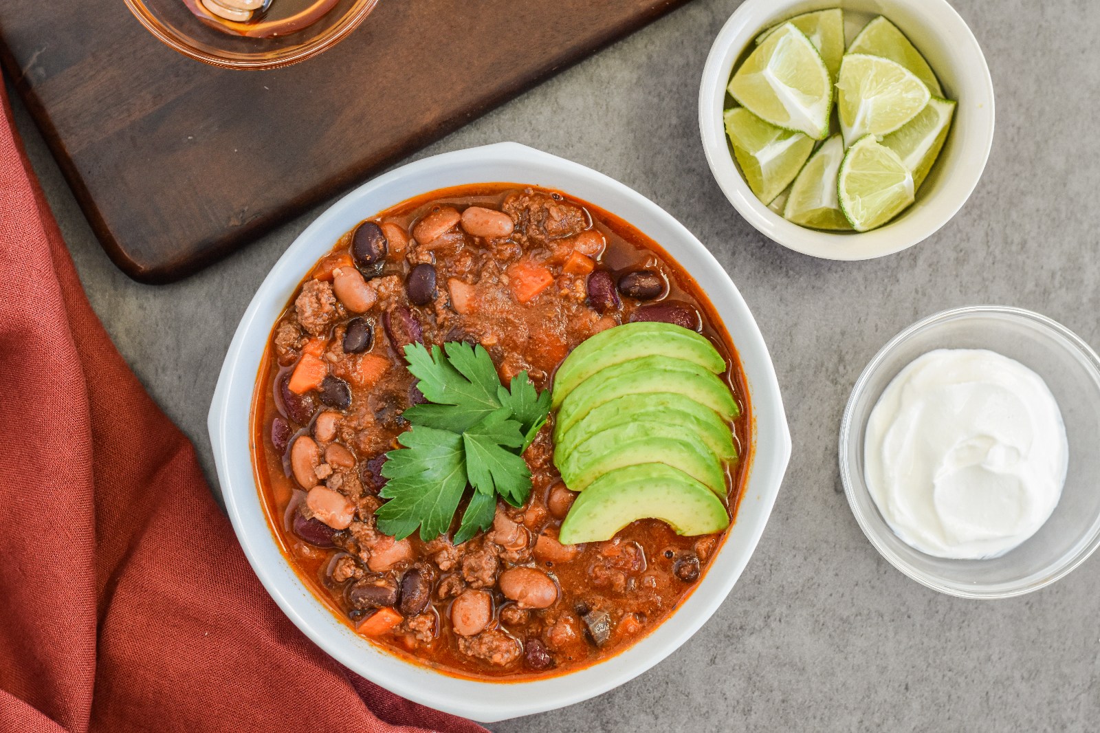 A white bowl filled with three bean chili and a bowl of sliced limes and a spoon.