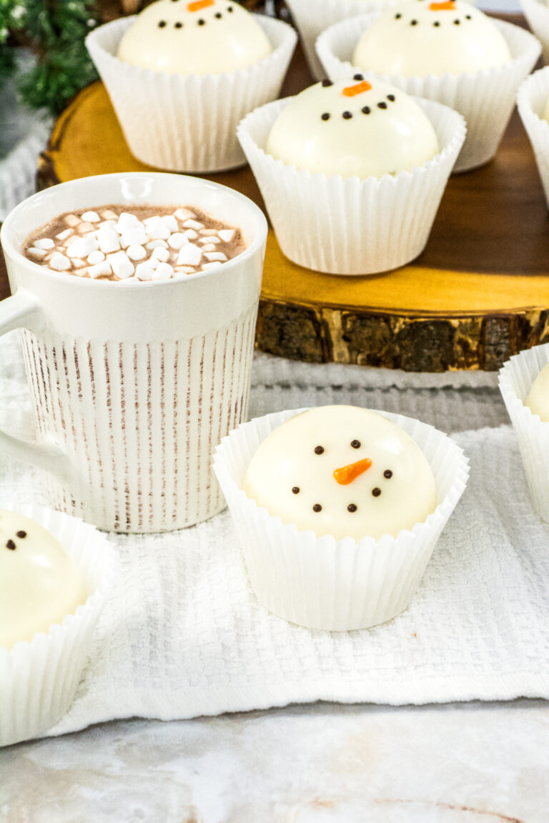 A white mug with marshmallows and hot chocolate with snowman hot chocolate bombs in candy wrappers.