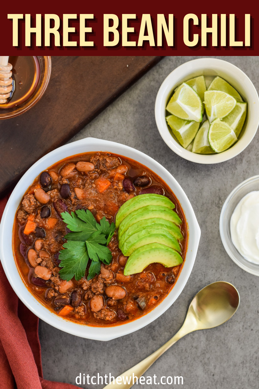 A white bowl of three bean chili with avocado slices and lime wedges.
