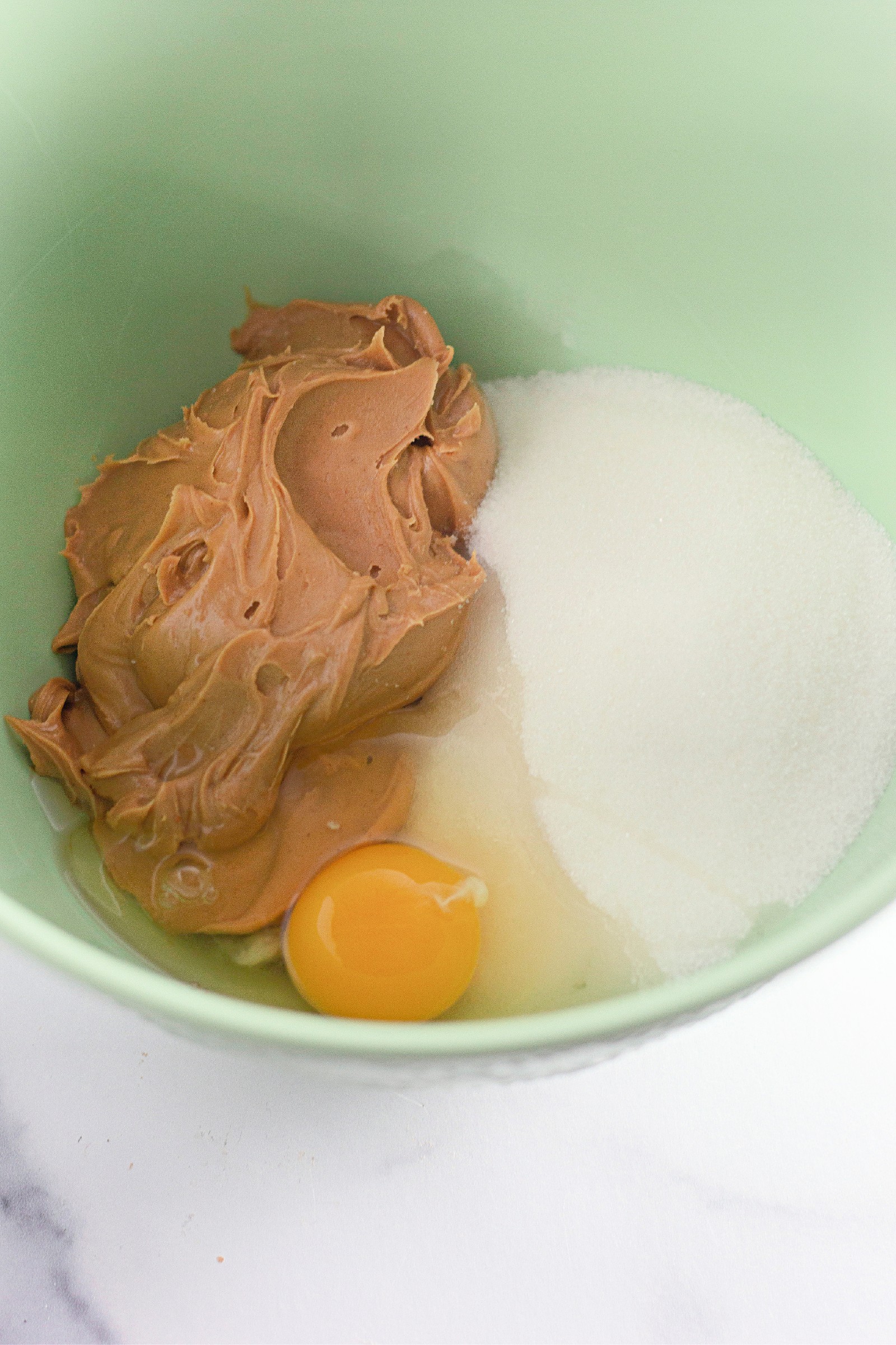 Peanut butter, sugar, and an egg in a green mixing bowl to make cookies.