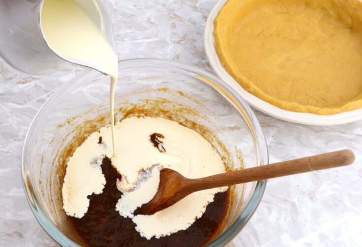 Heavy cream being poured into a glass bowl with pumpkin pie filling ingredients and a pie crust in the background.