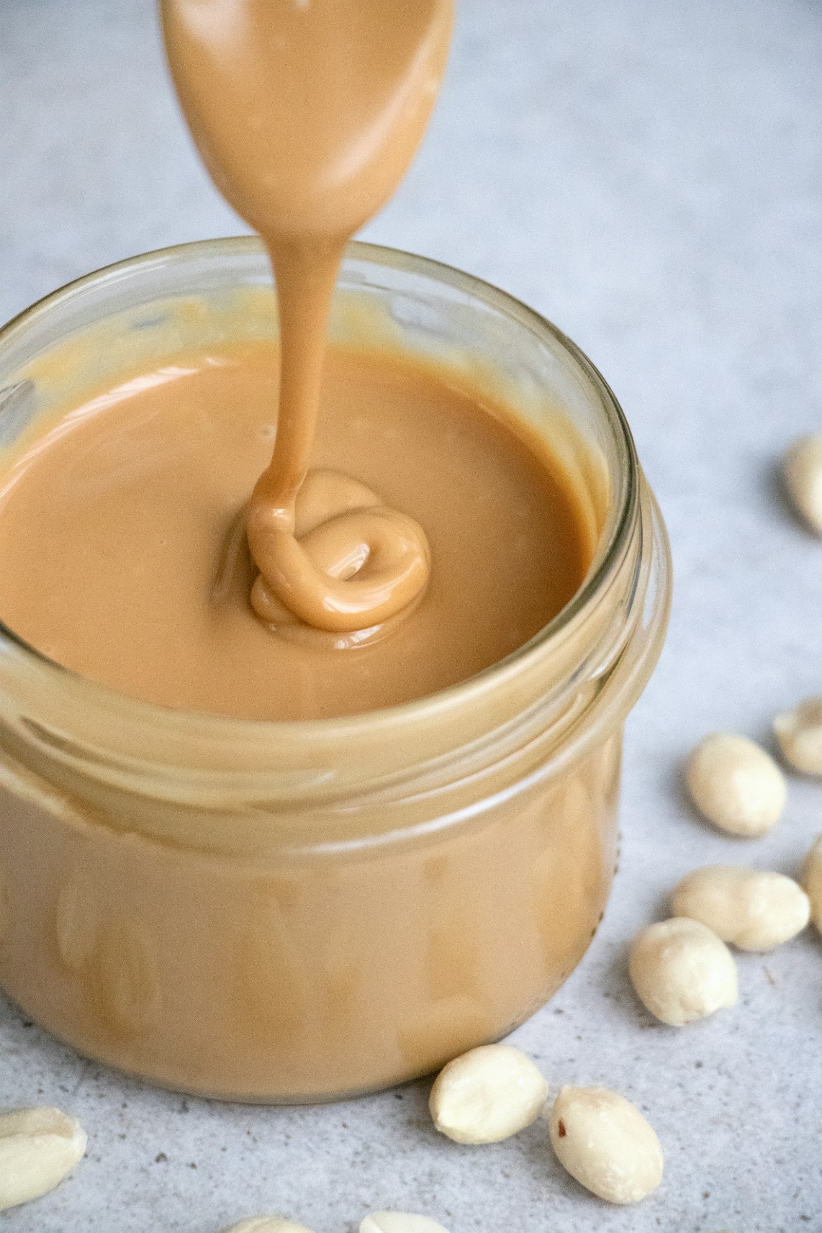 Peanut butter being poured into a glass jar with peanuts scattered around.
