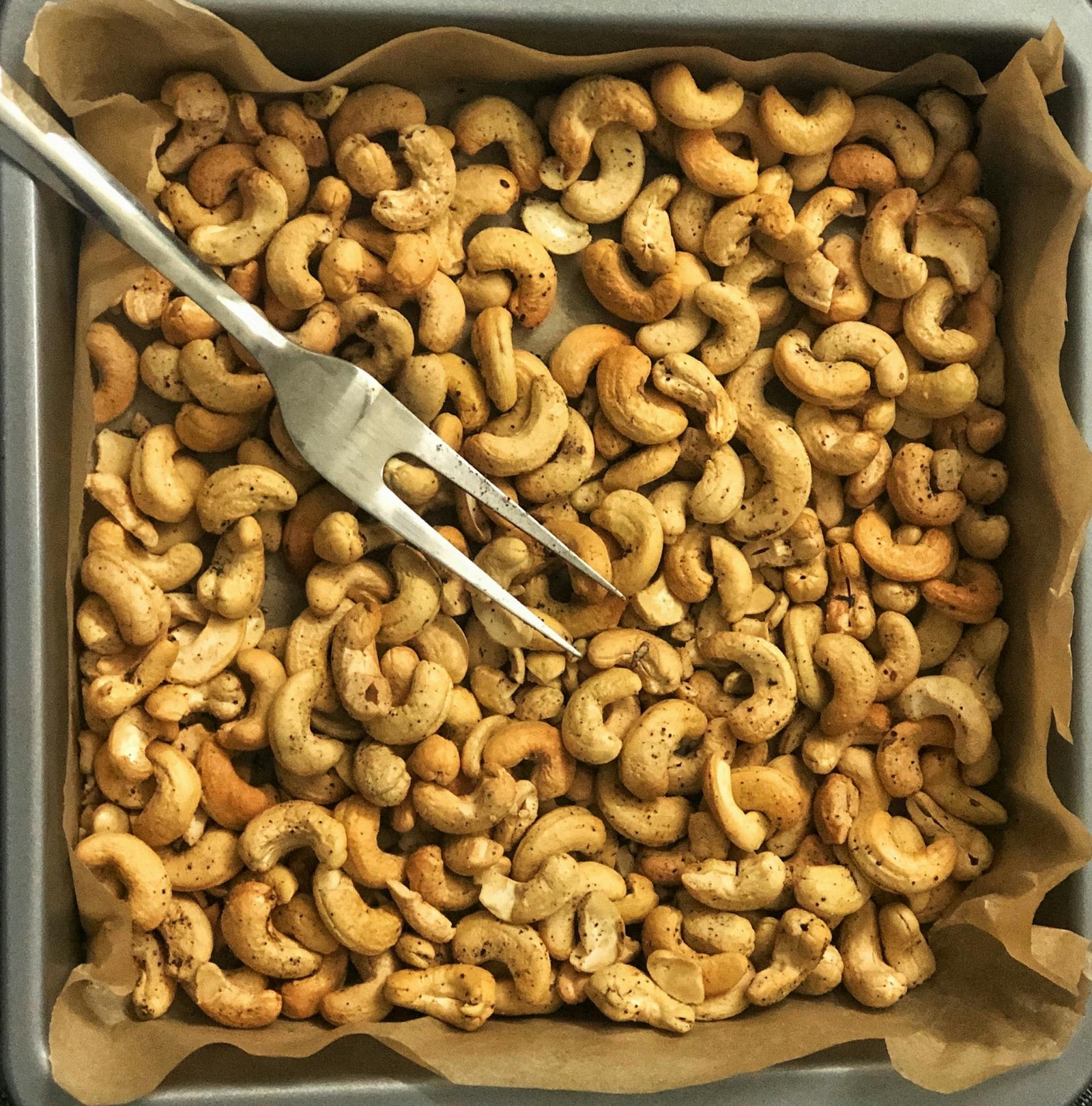A baking pan with cashews, parchment paper, and a large fork.