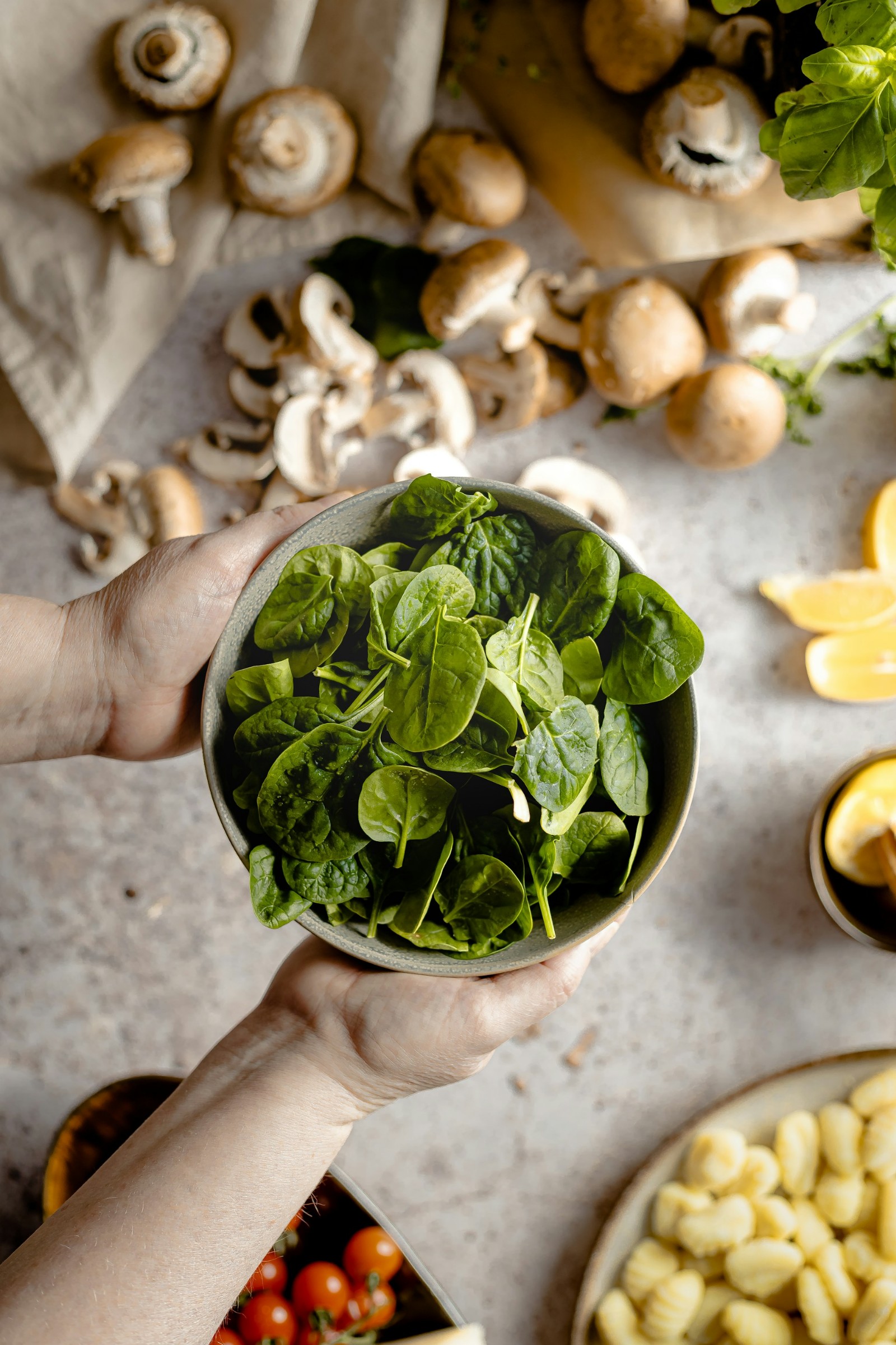 A bowl of spinach with hands holding it and mushrooms and other vegetables in the background.