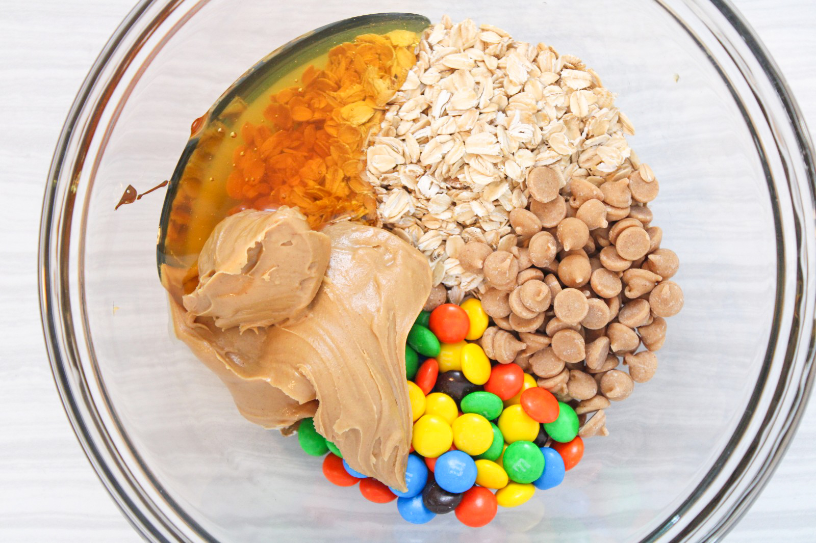 A glass bowl with oats, peanut butter chips, M&M's, peanut butter, and honey.