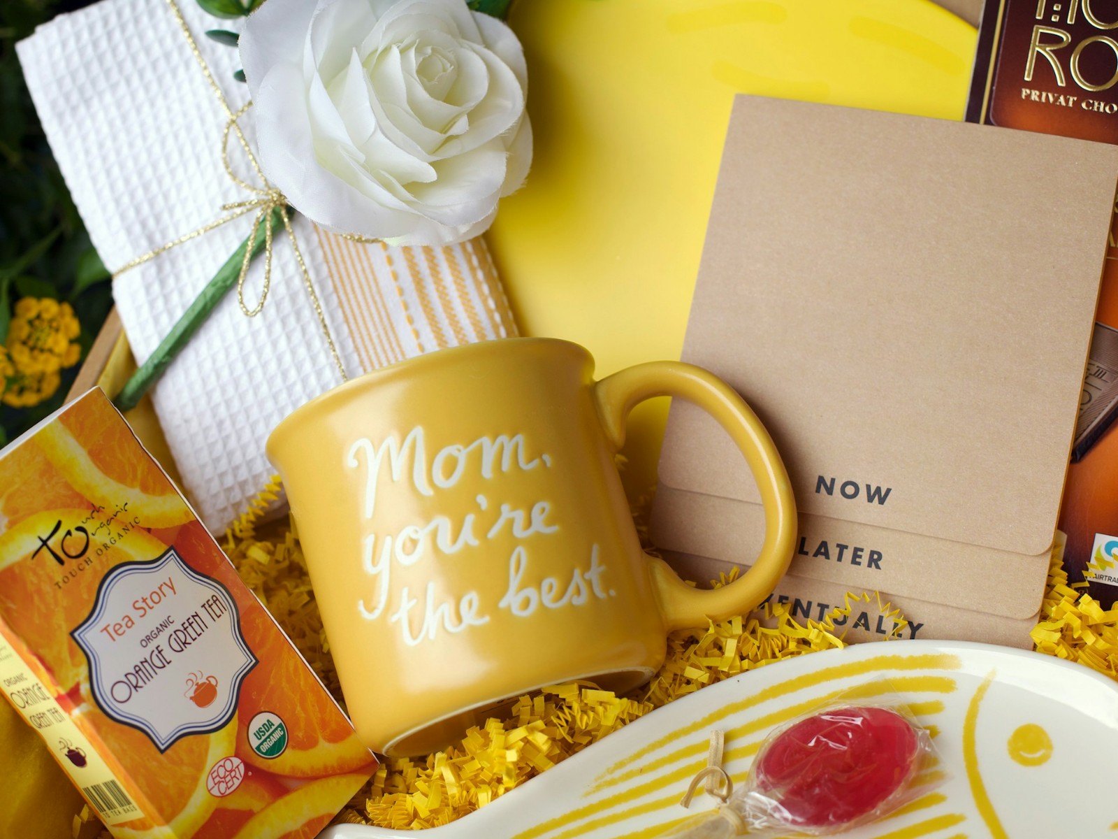 A basket filled with a yellow mug, chocolate, a white rose, and other little gifts.