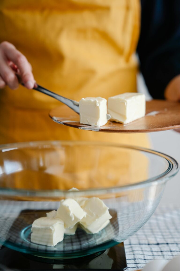 A woman, wearing a a yellow apron, adding butter to a glass bowl.