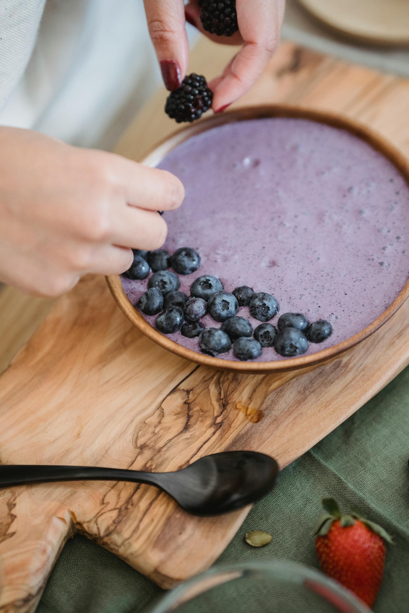 A blueberry smoothie bowl with a hand placing fruit on top of the bowl.