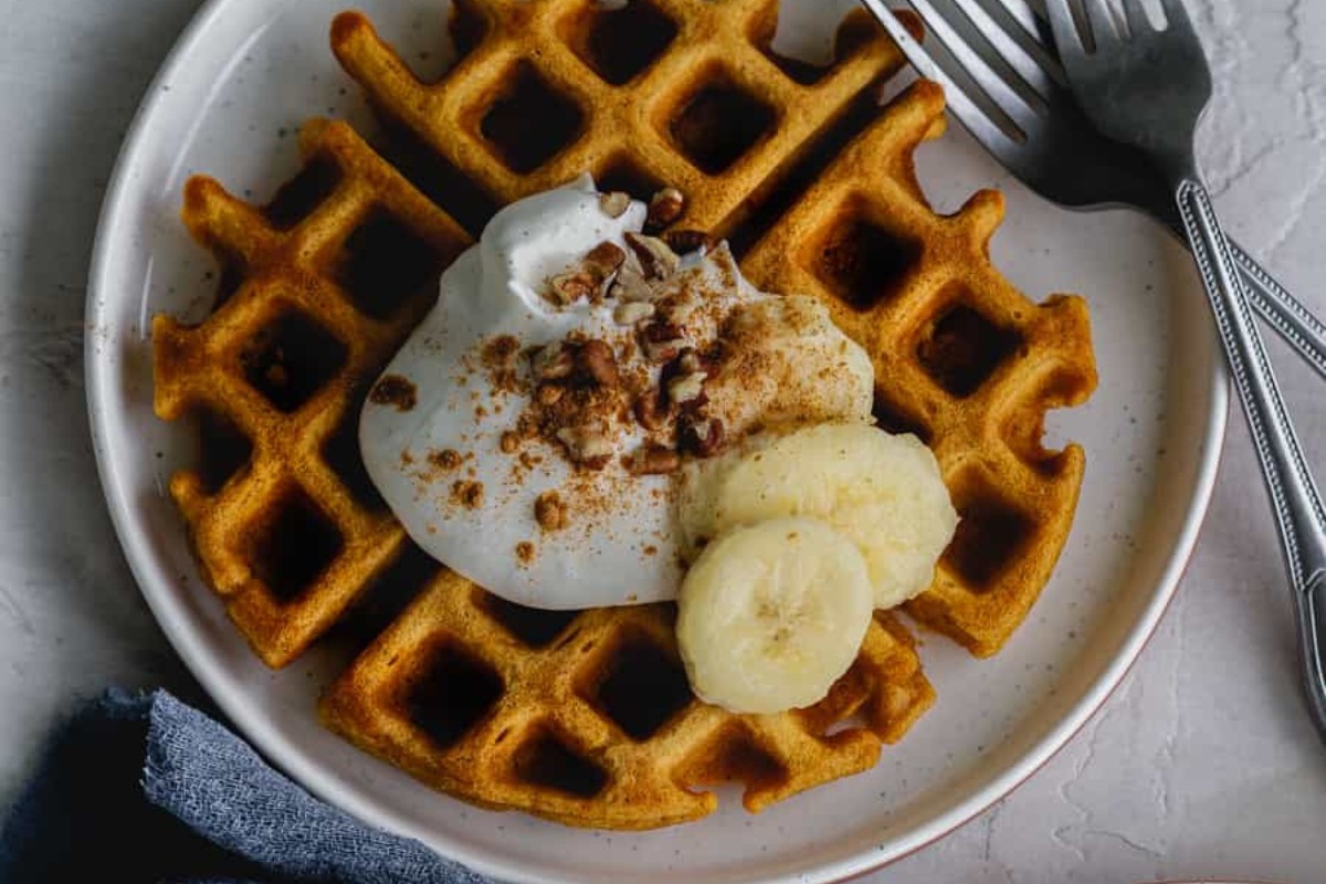 A white plate with one pumpkin waffle with sliced bananas and whipped cream and two forks.