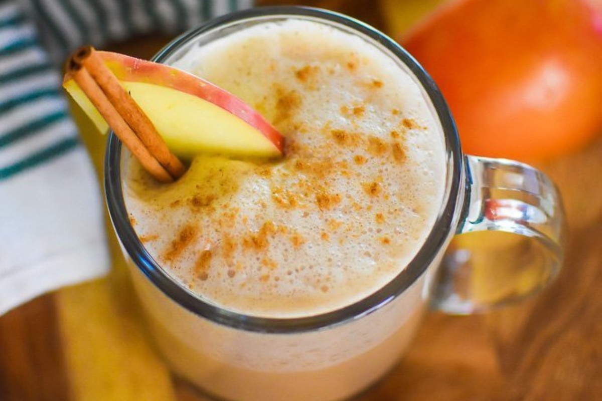 A glass mug filled with an apple butter chai latte with a few sliced apples.