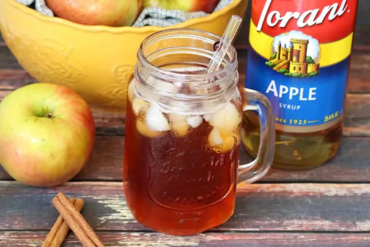 A glass filled with ice and spiced Apple Iced tea with an apple and cinnamon sticks and a bottle of apple syrup.